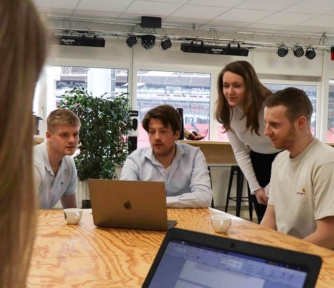 Vier groeileaders zitten en staan rond een tafel terwijl ze samen naar een laptop kijken tijdens een meeting, met koffiekopjes op tafel.