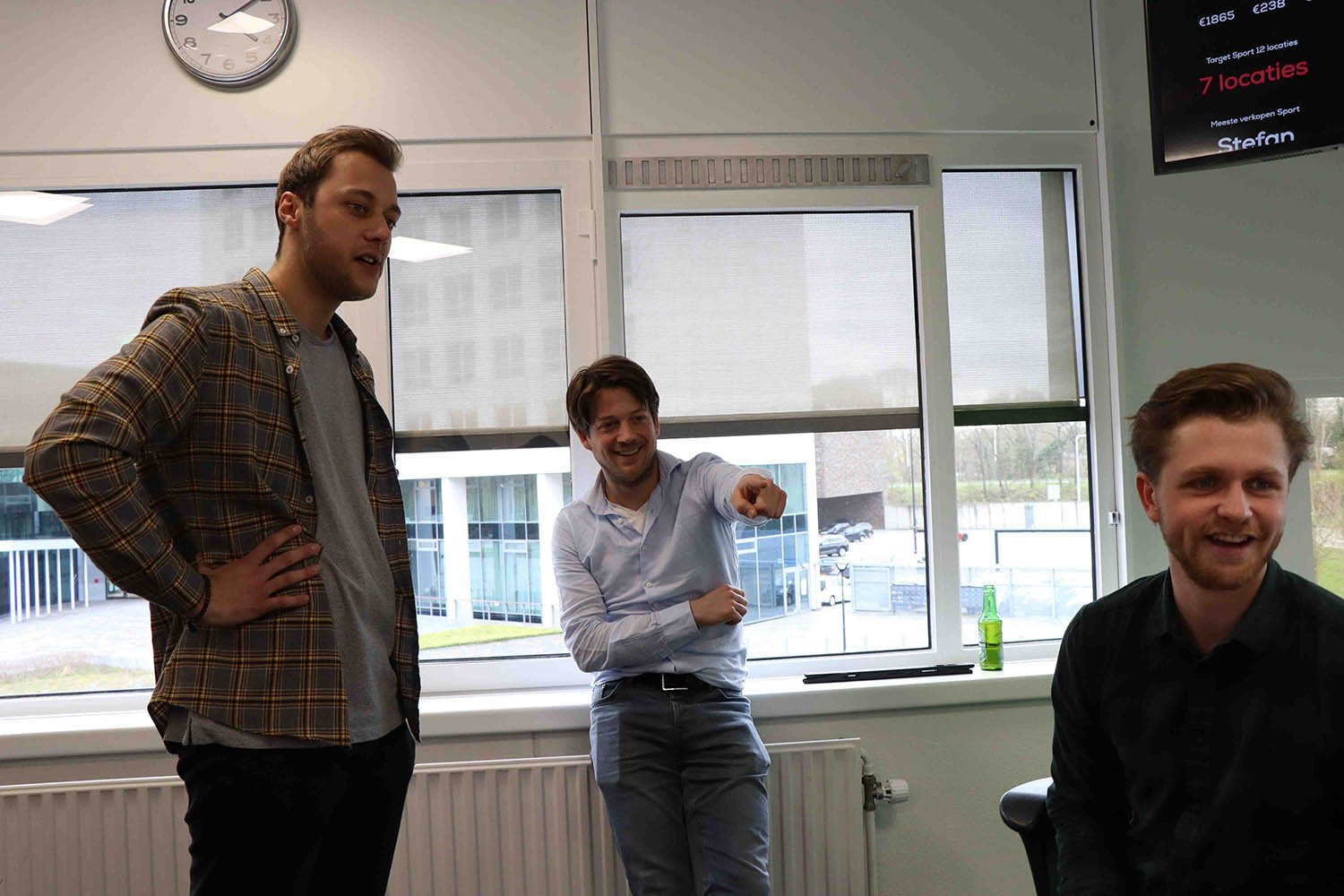 Drie groeileaders in een kantoorruimte praten en lachen met elkaar; één staat met hand in de zij, een ander wijst lachend naar voren, en de derde zit glimlachend op een stoel.