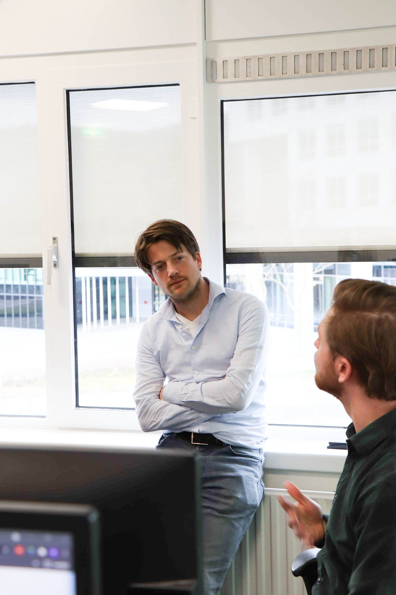 Twee groeileaders in gesprek in een kantoor; de een leunt met armen over elkaar tegen het raam, terwijl de ander op een stoel zit en praat.