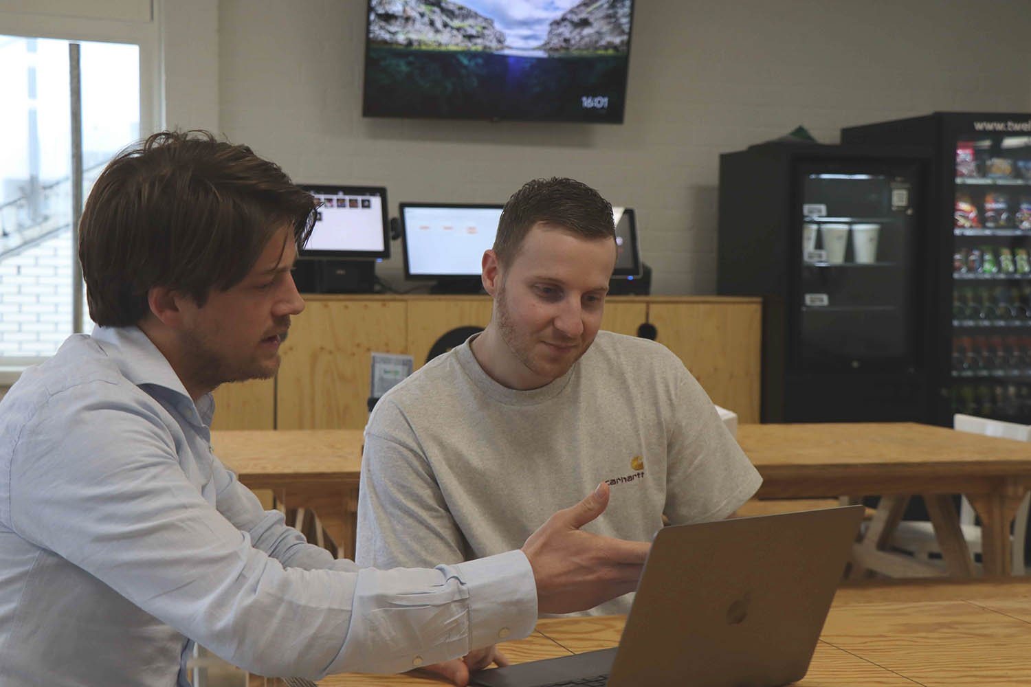 Twee groeileaders zitten samen aan een tafel in een kantine en kijken naar een laptop, terwijl één van hen iets uitlegt.