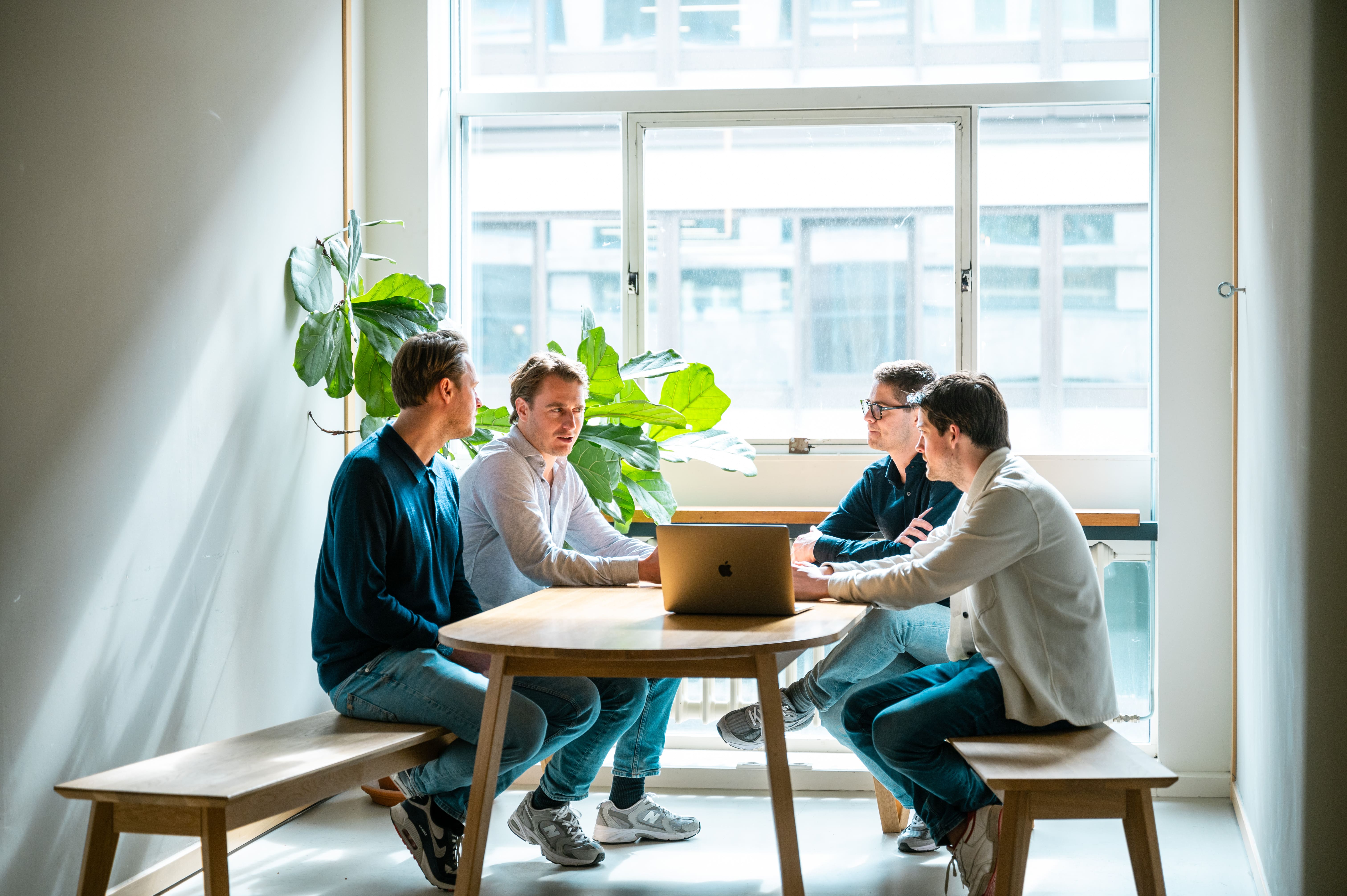 Vier groeileaders zitten en staan rond een tafel en kijken lachend naar een laptop tijdens een informele meeting.