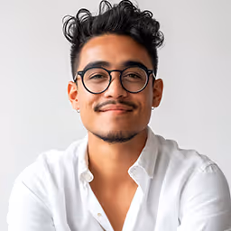 Smiling young man with curly hair and glasses wearing a white button-up shirt against a plain background.