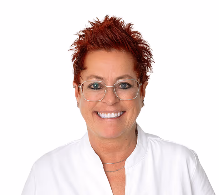 Porträt einer lächelnden Frau mit kurzen, roten, stacheligen Haaren und Brille, die eine weiße Bluse trägt.