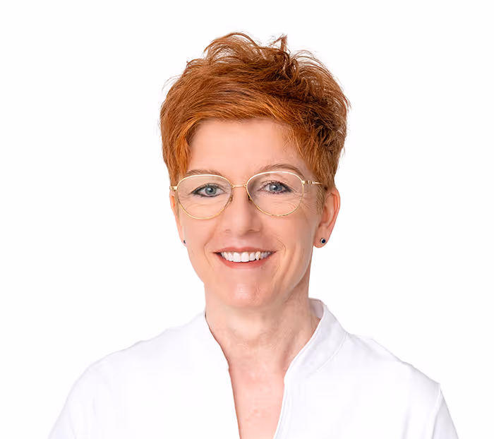 Ältere Frau mit kurzen roten Haaren, Brille und weißer Bluse lächelt vor weißem Hintergrund.