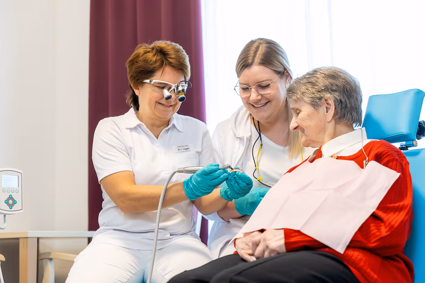 Zahnärztin behandelt lächelnde ältere Patientin, während Helferin assistiert im Behandlungszimmer.