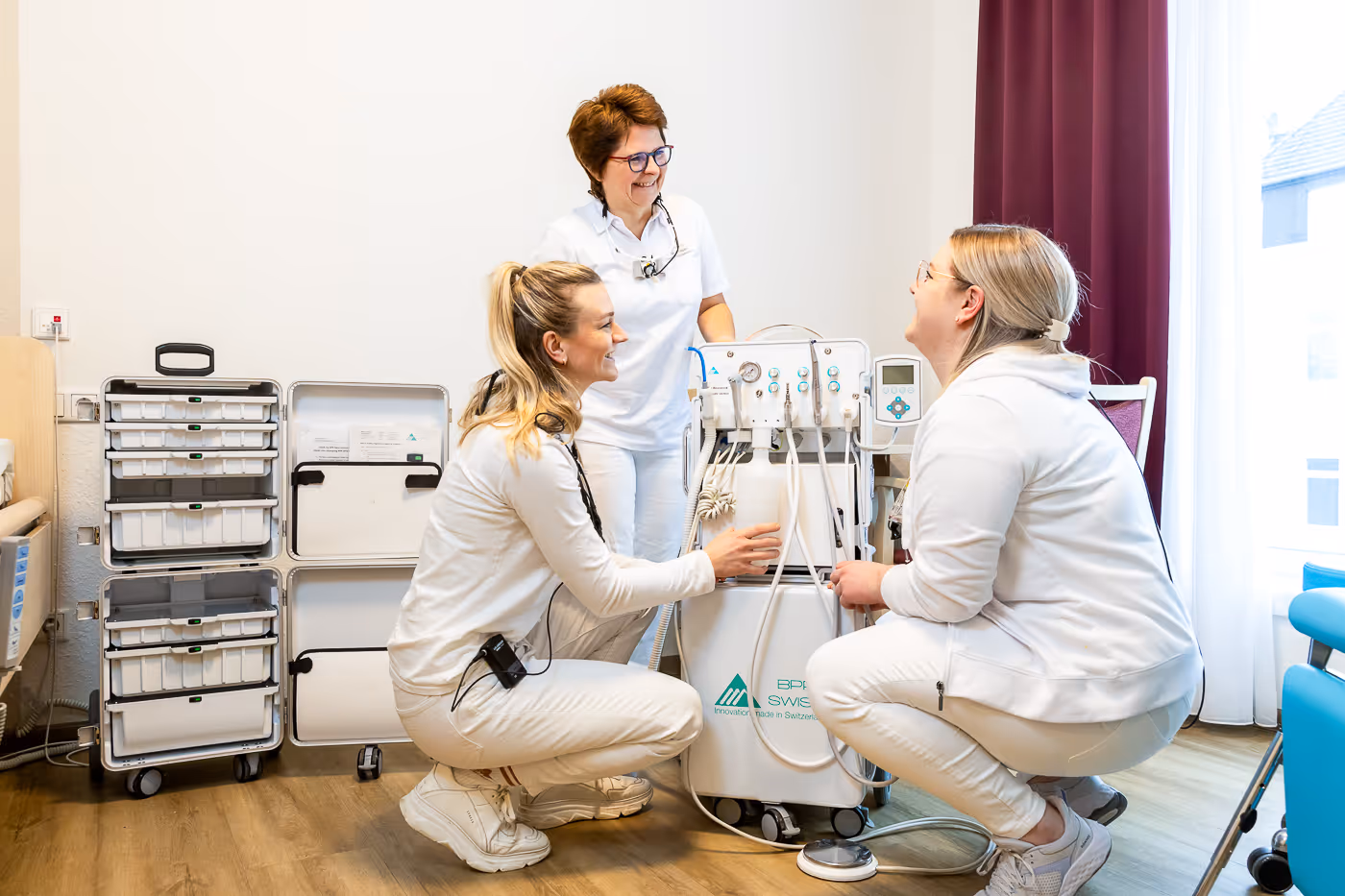 Drei Frauen in medizinischer Arbeitskleidung lächeln und benutzen ein medizinisches Gerät mit Schläuchen in einer hellen Behandlungszimmerumgebung.