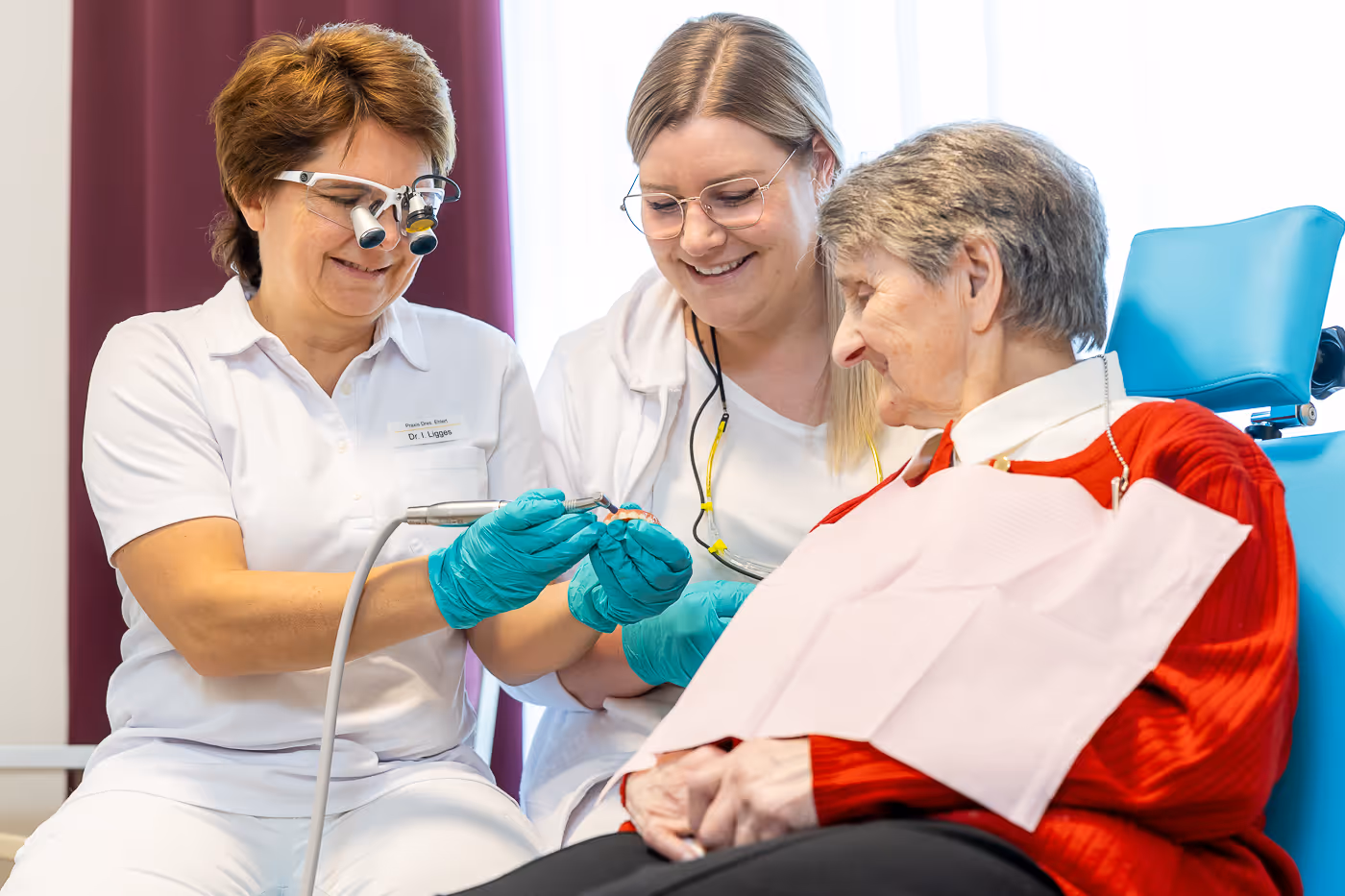 Zahnärztin und Assistentin lächeln, während sie ein Zahnmodell für eine ältere Patientin in rotem Pullover erklären.