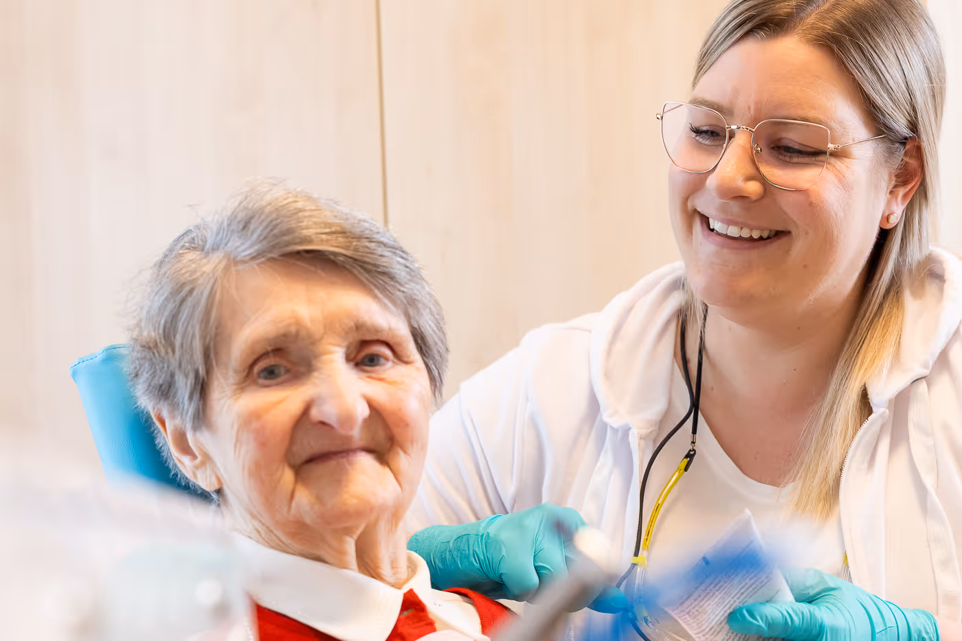 Ältere Frau lächelt leicht, während eine Pflegekraft mit blauen Handschuhen sie fürsorglich betreut.