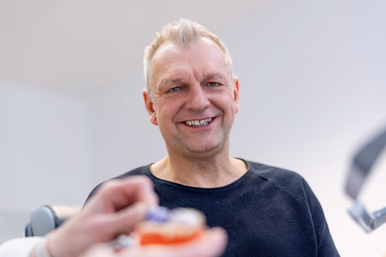 Mann mittleren Alters lächelt in einer Zahnarztpraxis, während ihm ein Zahnarzt ein Zahnmodell zeigt.