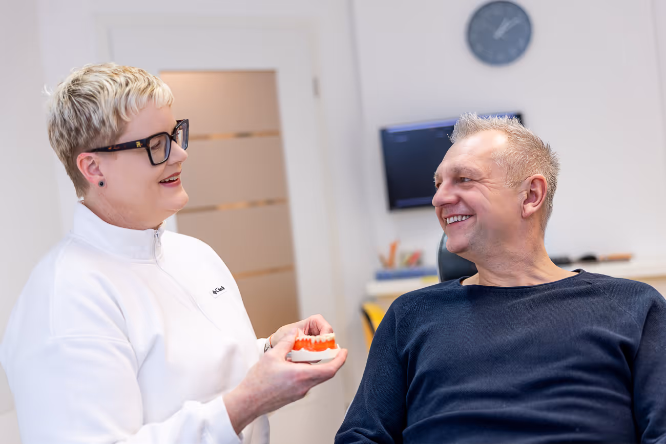 Mitarbeiterin hält ein Zahnmodell und spricht mit einem lächelnden männlichen Patienten in einer Praxis.
