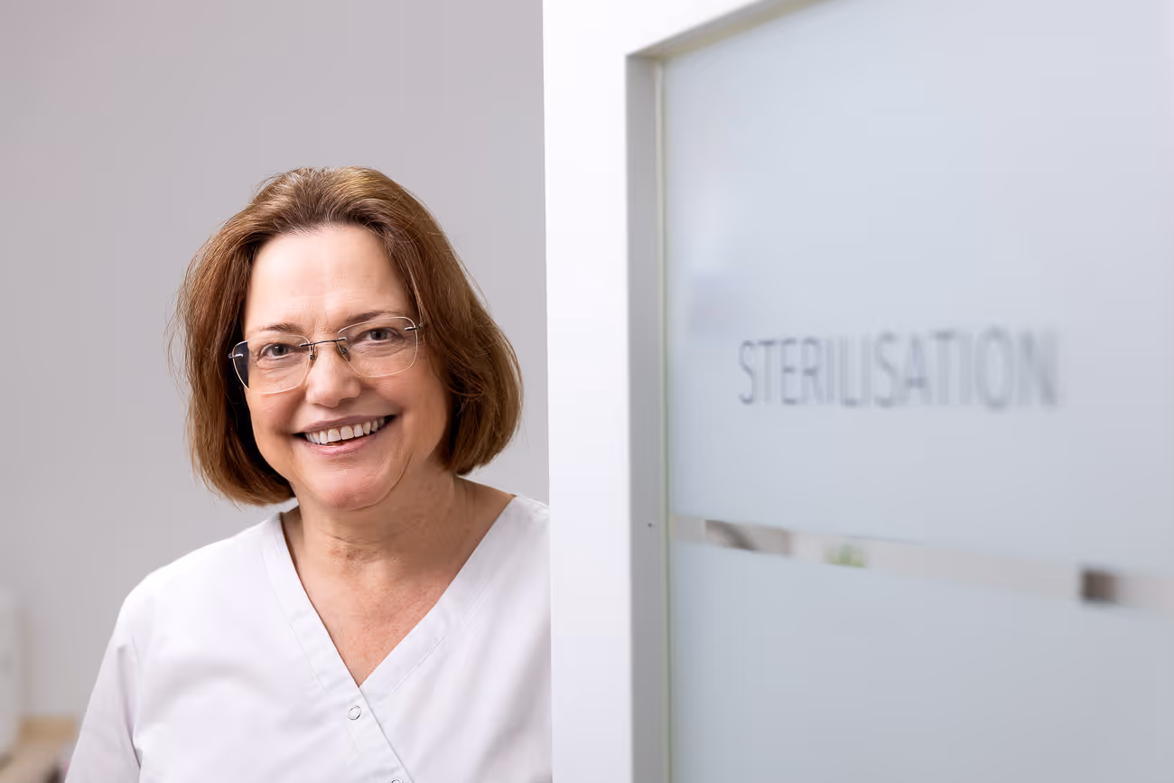 Ältere Frau in weißer Arbeitskleidung lächelt vor einer Glastür mit der Aufschrift Sterilisation.
