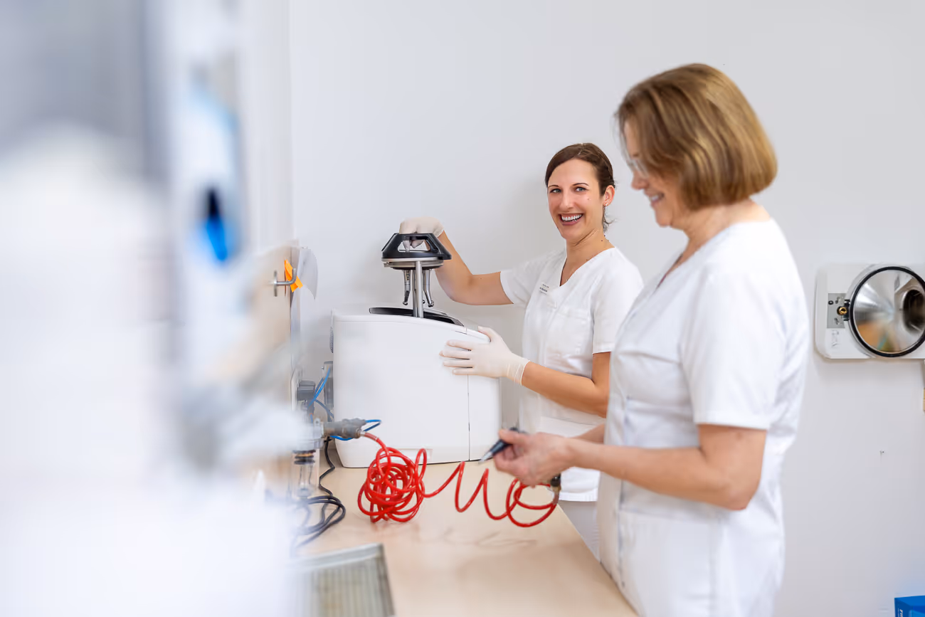 Zwei Frauen in weißen medizinischen Kitteln arbeiten gemeinsam mit einem Laborgerät und einem roten Kabel in einem hellen Labor.