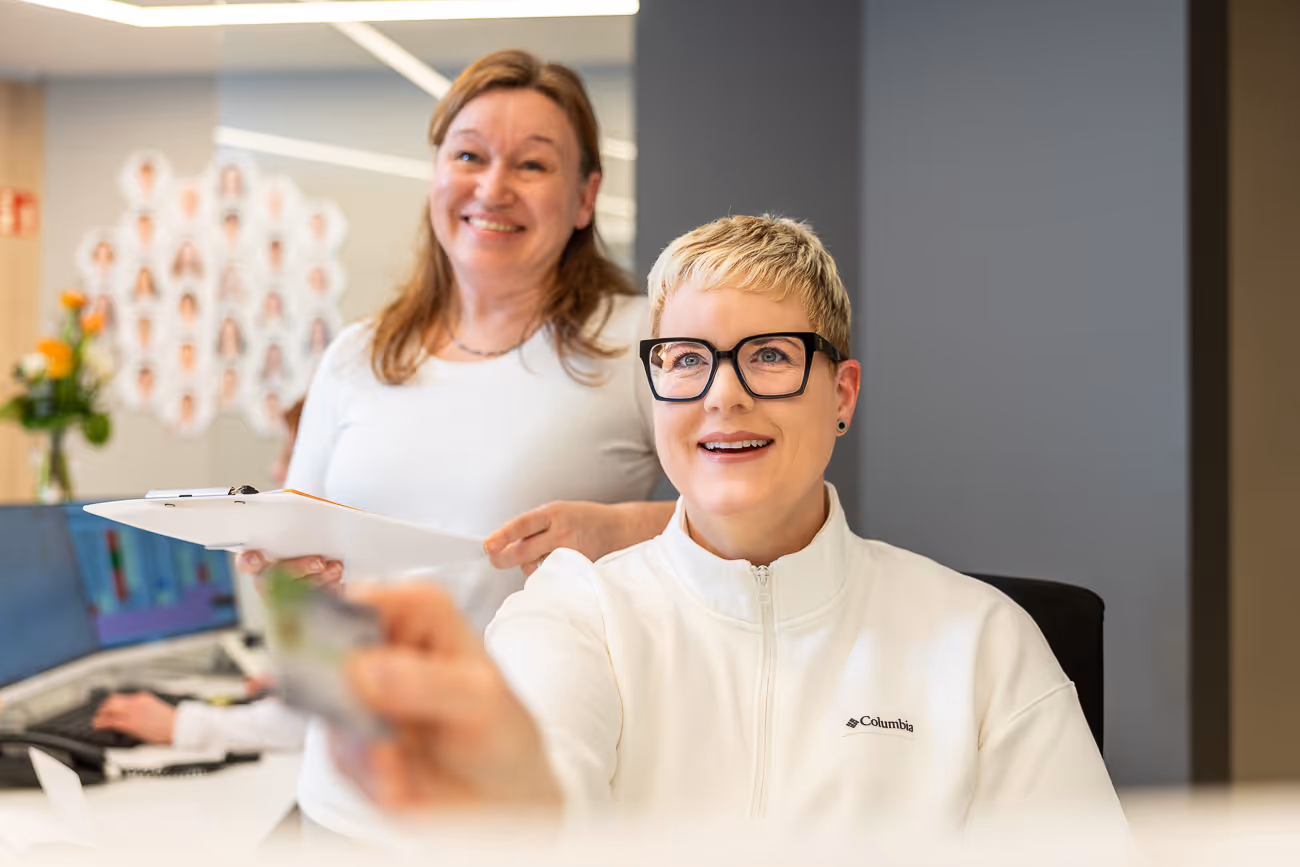 Lächelnde Frau mit kurzen blonden Haaren und Brille reicht eine Karte nach vorne, während eine andere Frau mit braunen Haaren und Klemmbrett im Hintergrund steht.
