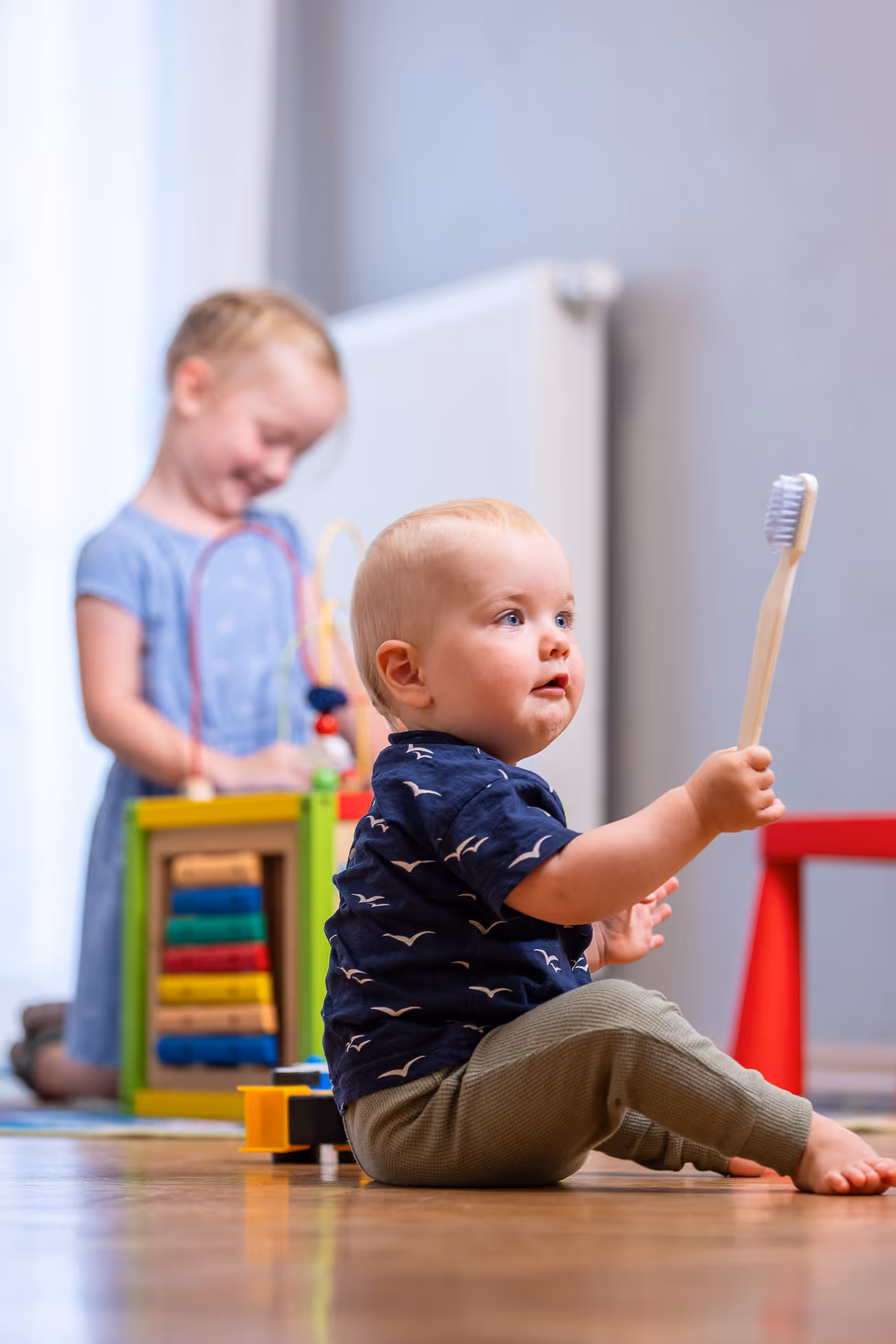 Kleinkind sitzt auf dem Boden und hält eine Zahnbürste, im Hintergrund spielt ein anderes Kind mit einem bunten Spielzeug.