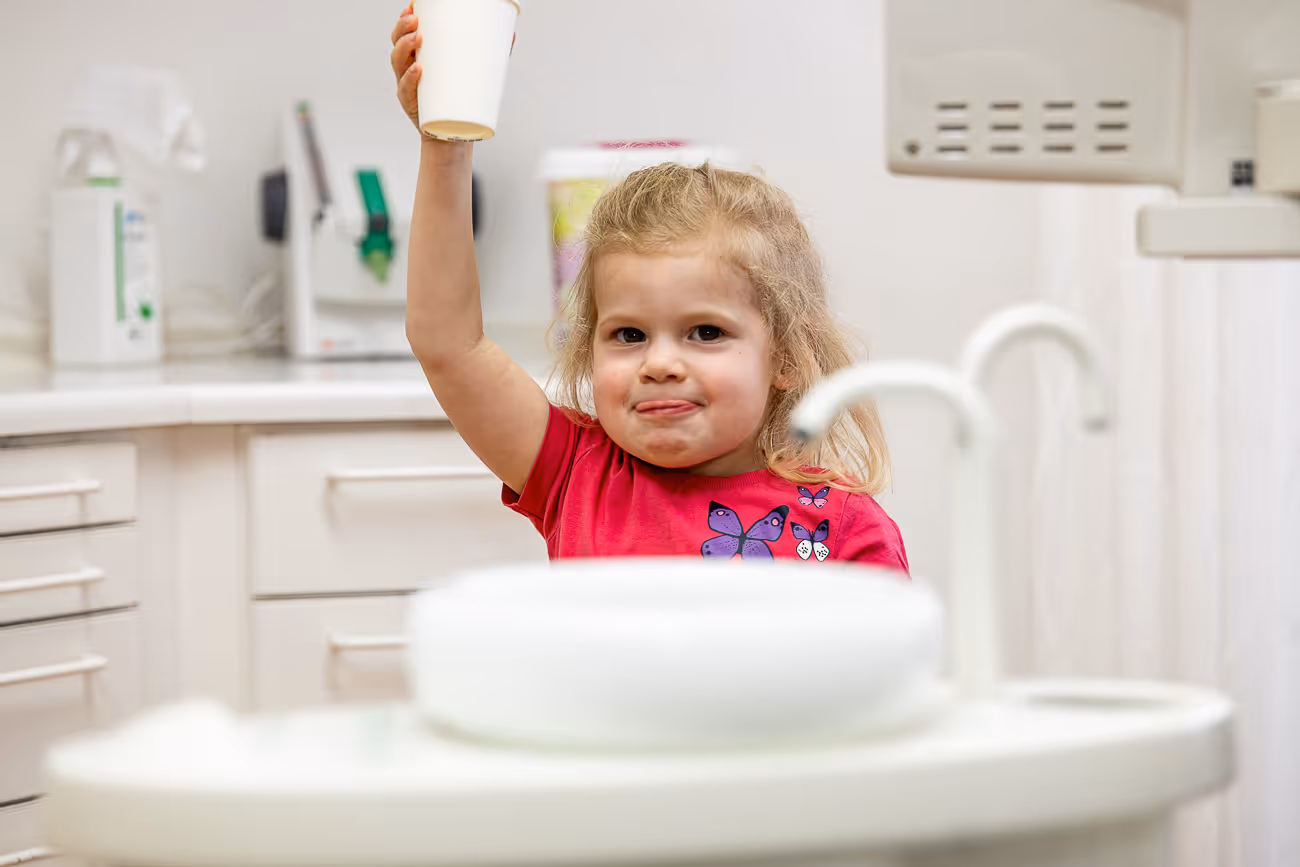 Kleines Mädchen in einer Zahnarztpraxis hält einen Becher hoch und blickt in die Kamera.
