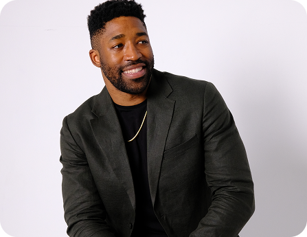 Meech smiling wearing a dark blazer and black shirt with a gold chain necklace against a light background.