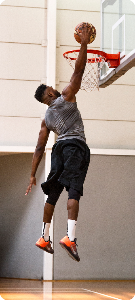 Meech in gray tank top and black shorts jumping to dunk a basketball in an indoor court.