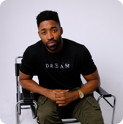 Meech with short curly hair and beard wearing a black t-shirt with the word 'DREAM' sitting on a metal-frame chair with hands clasped.