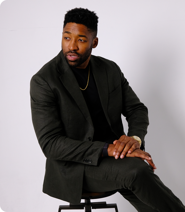 Meech with black hair and beard wearing a dark suit and gold chain sitting on a stool against a white background.