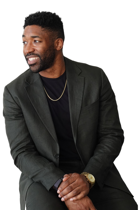 Meech Robinson (Meech) smiling wearing a dark suit jacket, black shirt, gold chain, and gold watch sitting with hands clasped.