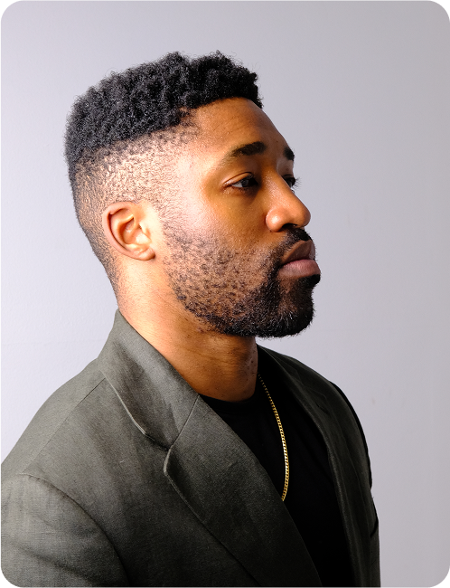 Side profile of Meech Robinson wearing a dark blazer and a gold chain against a plain light background.