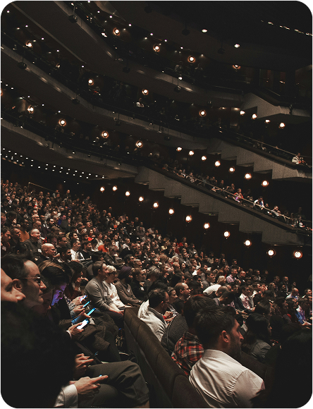 Large audience seated in a multi-tiered theater facing the stage attentively.