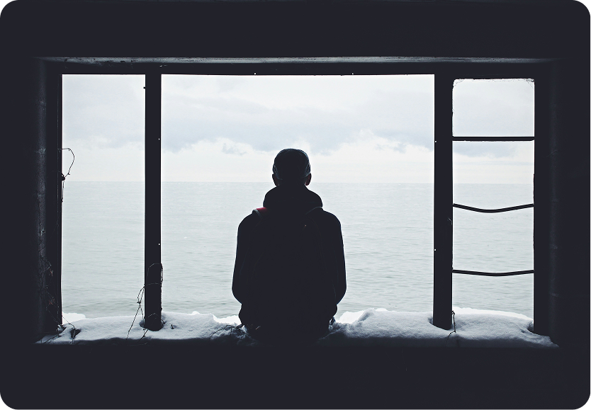Silhouette of a person sitting and looking out at a calm sea through a large window frame.