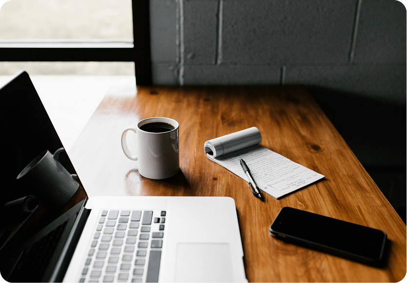 Wooden desk with an open laptop, a white coffee mug, a notepad with handwritten notes and a pen, and a smartphone.