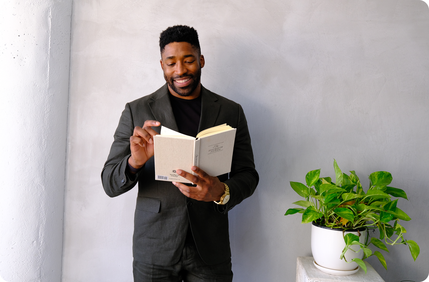 Meech Robinson (Meech) smiling in a dark suit reading a beige book near a potted green plant on a white stool against a gray wall.
