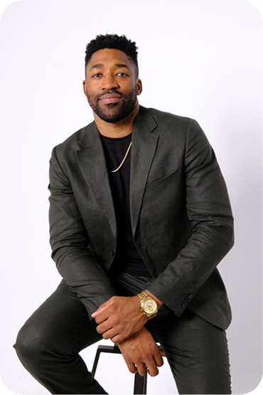 Meech Robinson (Meech) wearing a dark suit sitting on a stool against a plain white background, with a gold watch and chain.