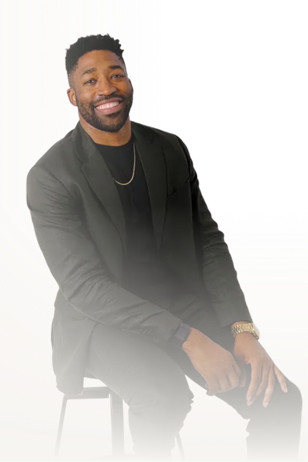 Meech smiling with a beard wearing a black blazer and gold jewelry sitting on a stool against a white background.