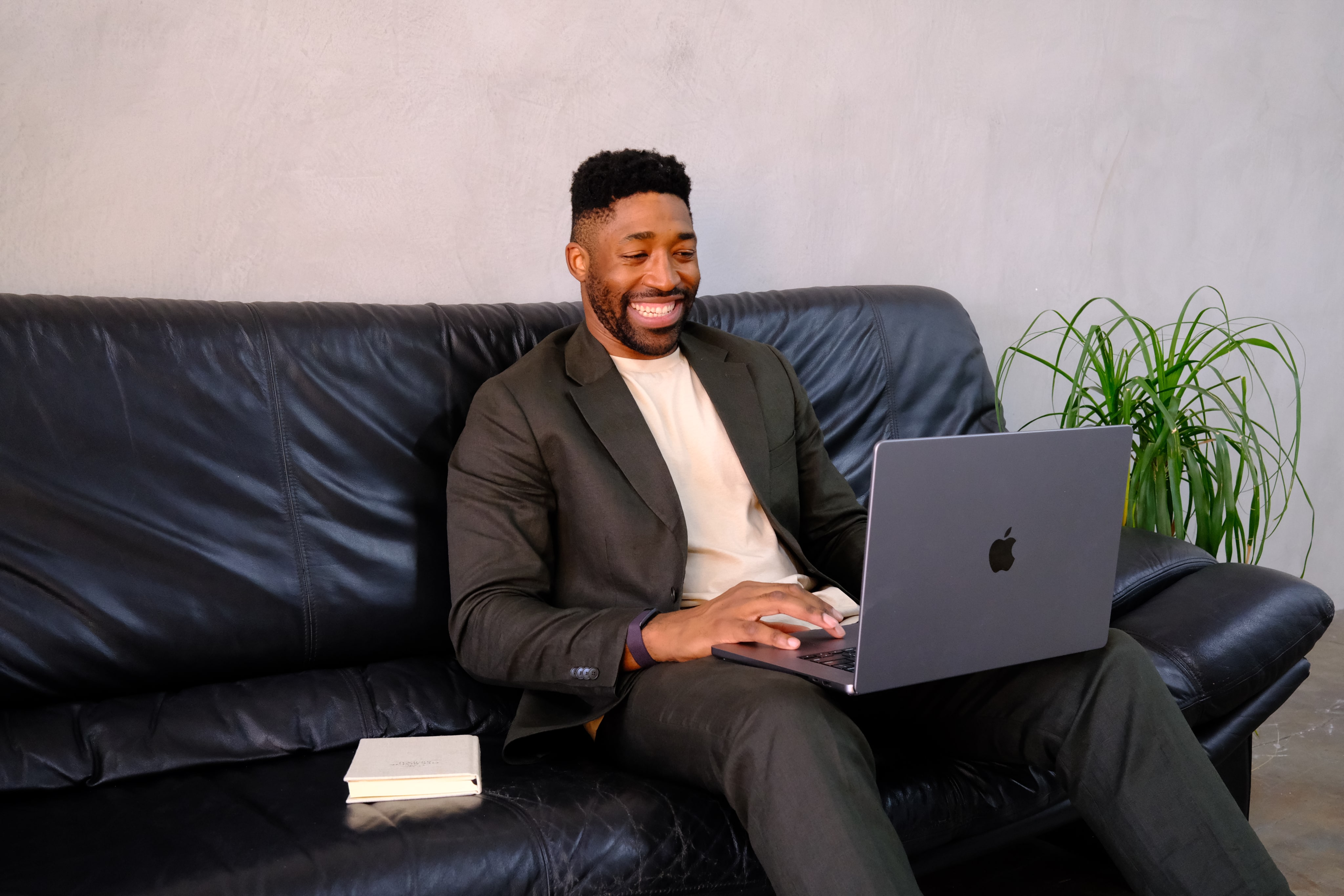 Meech smiling in a dark suit sitting on a black leather couch using a laptop with a small potted plant in the background.