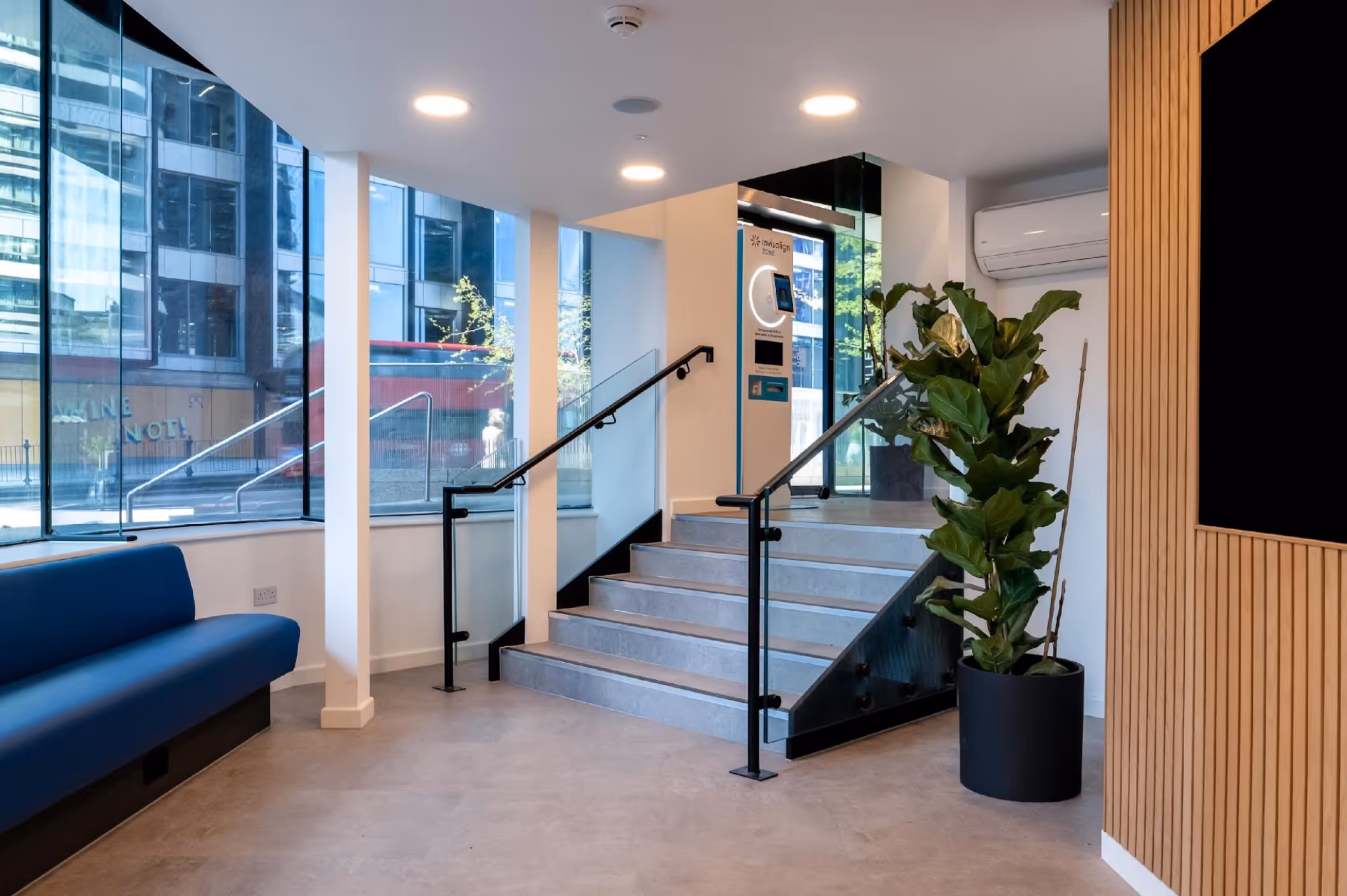 Modern interior with concrete stairs, black metal railings, large windows, blue bench seating, and a large potted plant.