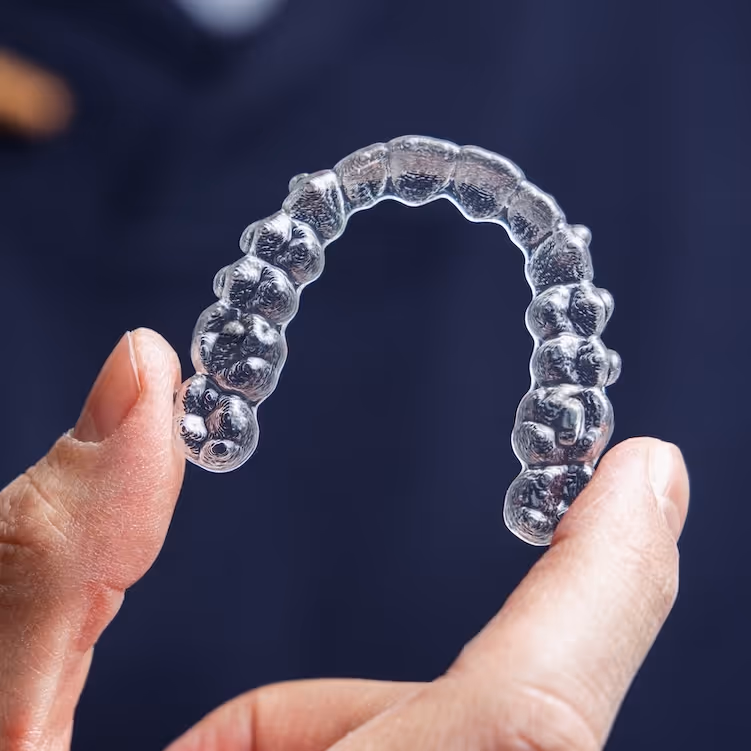 Hand holding a clear dental retainer against a dark blue background.