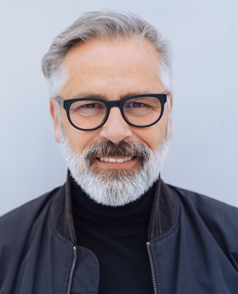 Smiling middle-aged man with gray hair, beard, and black glasses wearing a black jacket and turtleneck.