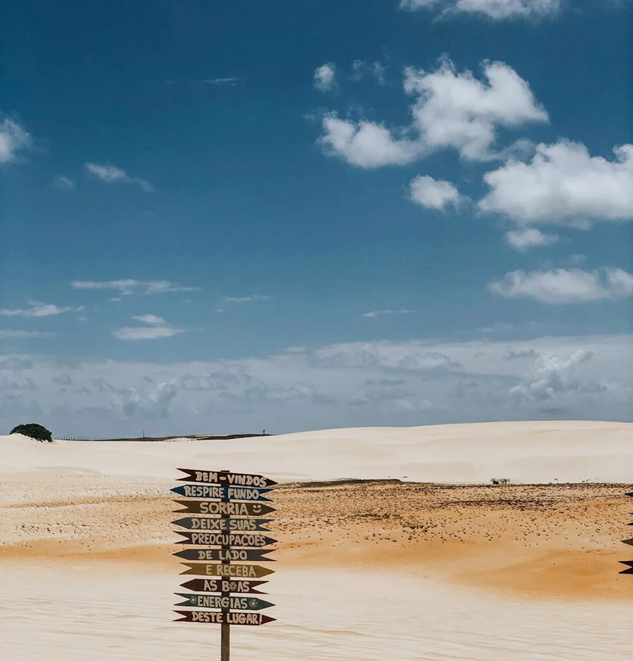 Wooden signpost with Portuguese welcome message in a sandy desert landscape under a partly cloudy sky.