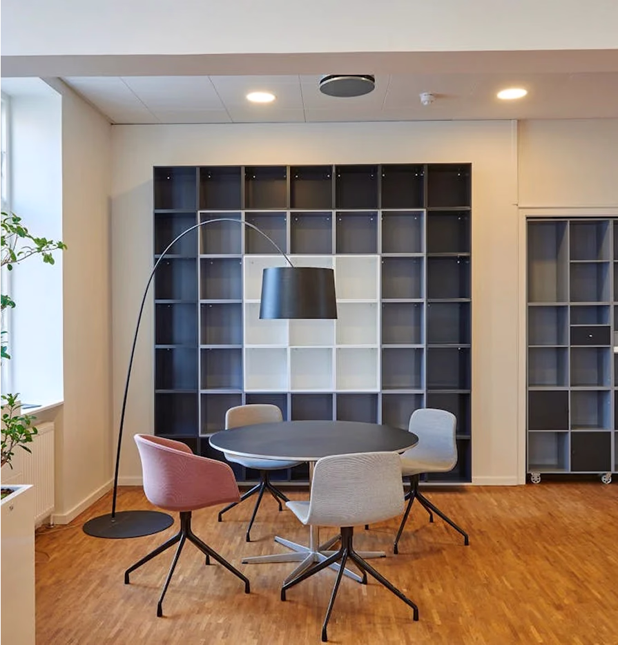 Modern meeting area with a round black table, four swivel chairs in neutral and pink colors, and a large arc floor lamp against a wall of empty square shelves.