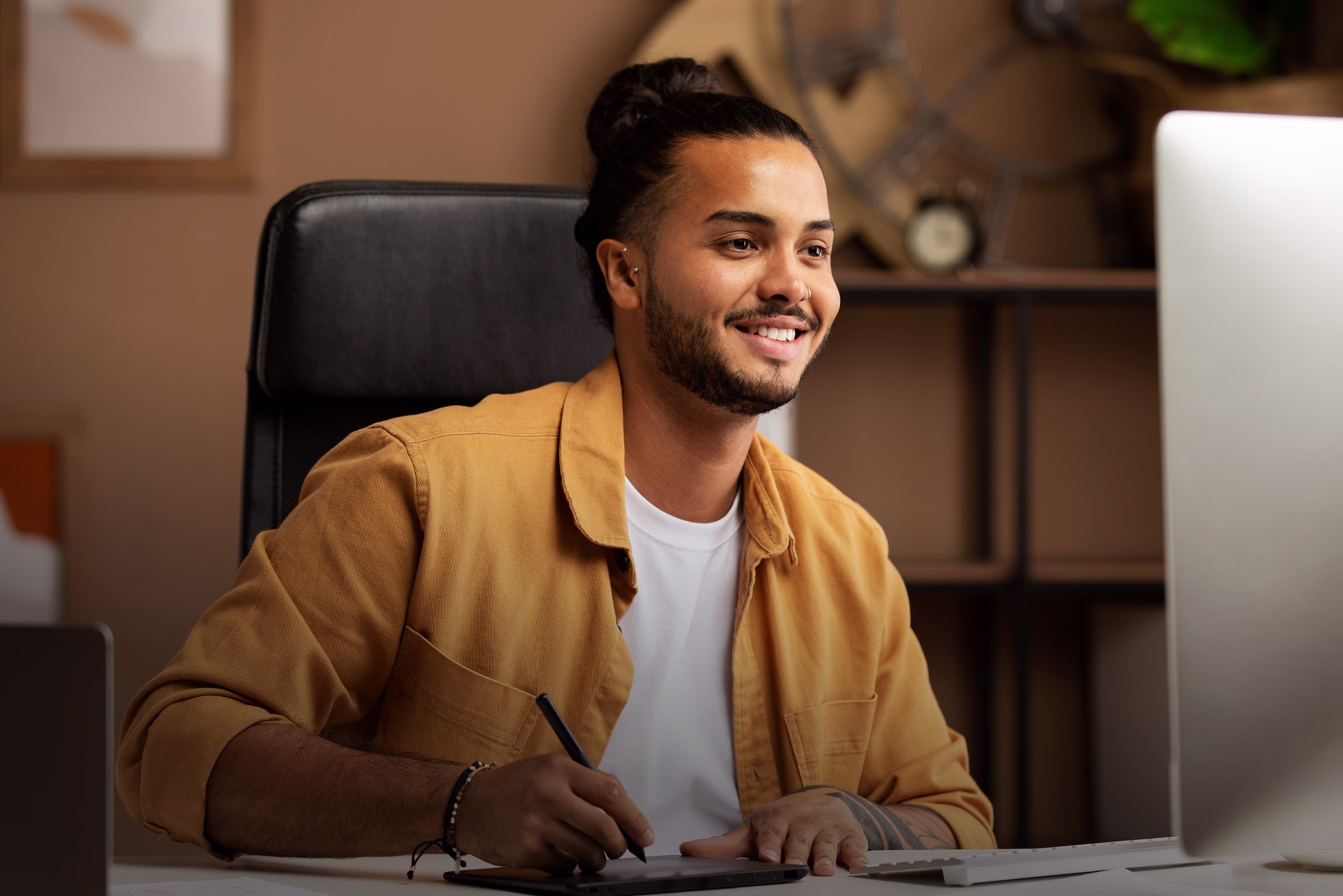Smiling man with a beard and man bun using a digital drawing tablet at a desk with a computer.