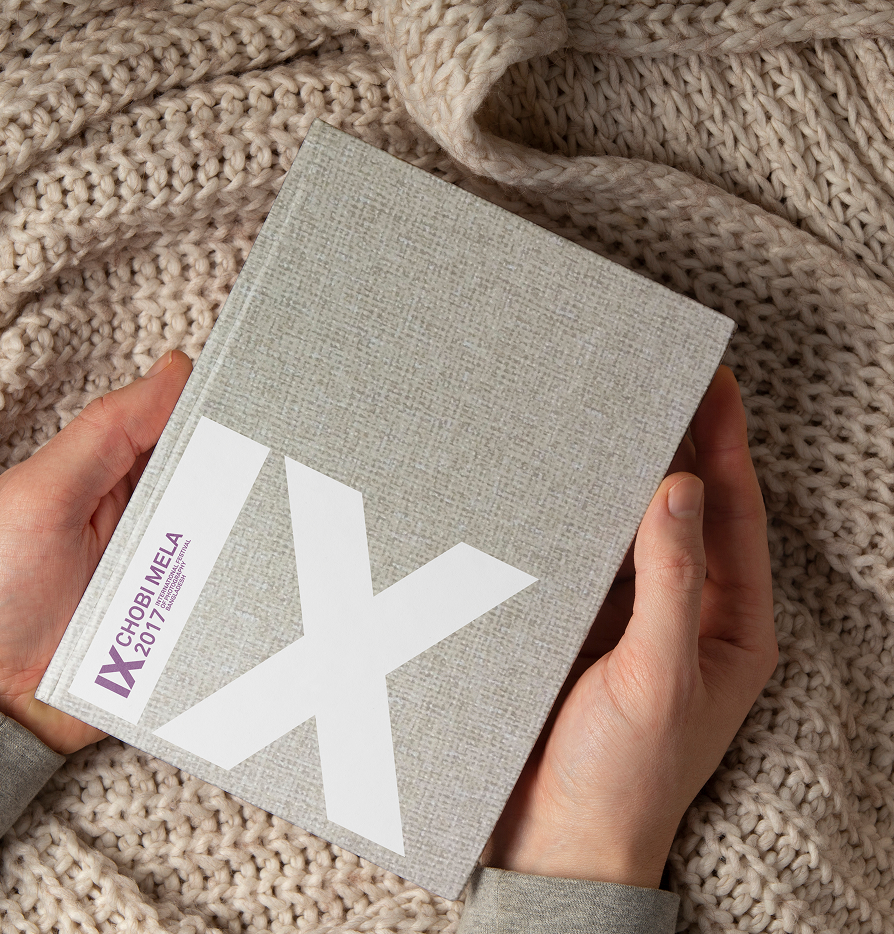 A person holding a beige book titled 'IX Chobi Mela 2017' against a knitted cream-colored blanket.