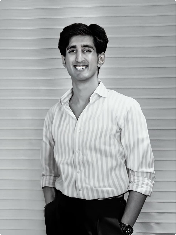 Young man smiling and posing with hands in pockets, wearing a striped shirt and dark pants against a light horizontal panel background.