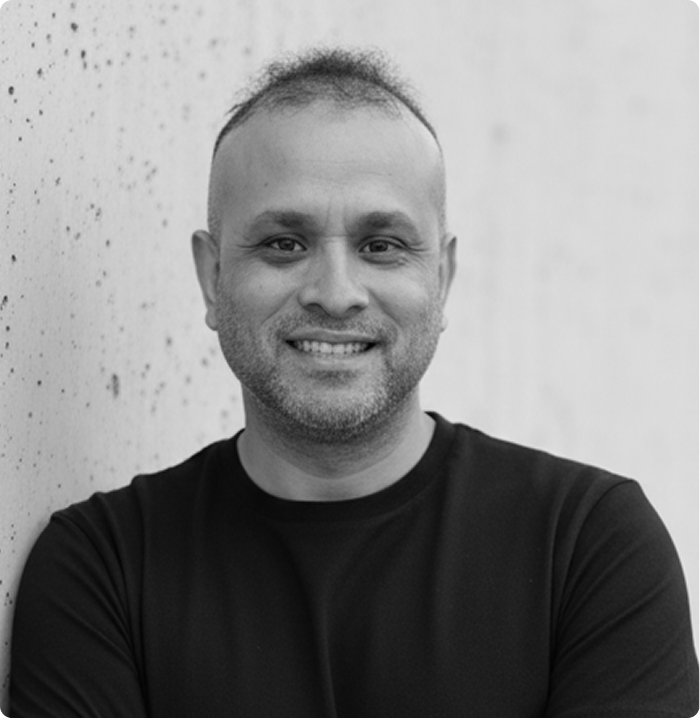 Smiling man with short hair and beard wearing a black shirt, leaning against a textured wall.