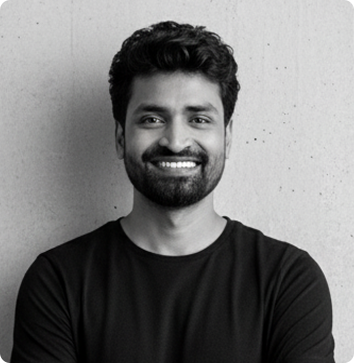 Smiling man with dark curly hair and beard wearing a black shirt standing against a light textured wall.