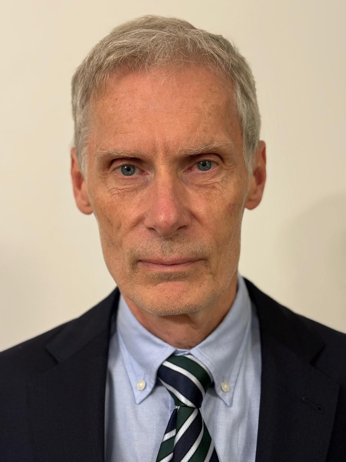 Portrait of an older man with gray hair wearing a suit, light blue shirt, and striped tie against a plain background.
