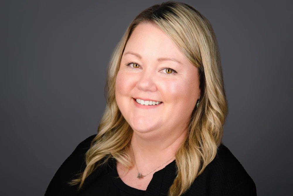 Smiling woman with blonde wavy hair wearing a black top against a gray background.