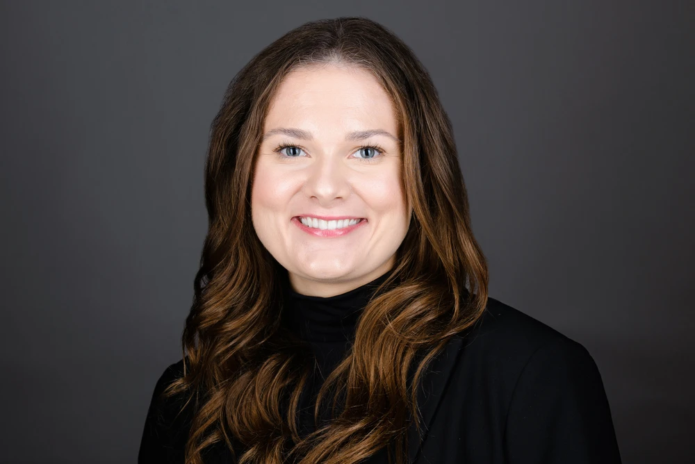 Smiling woman with long wavy brown hair wearing a black turtleneck and blazer against a gray background.