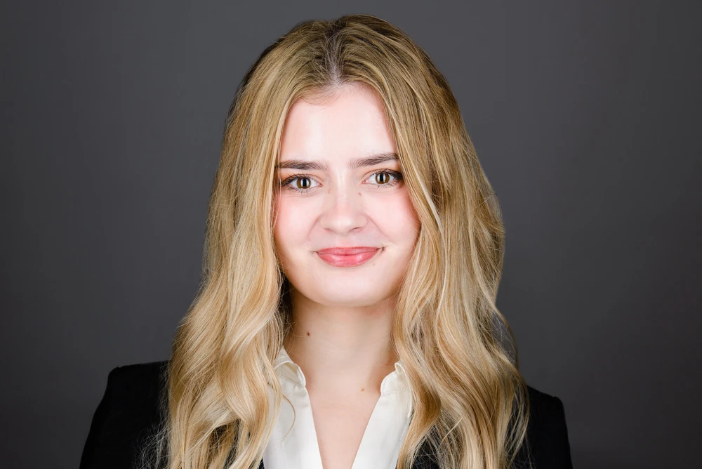 Smiling young woman with long blonde hair wearing a white blouse and black blazer against a gray background.