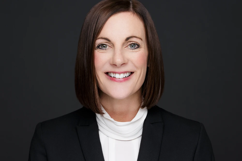 Smiling woman with straight shoulder-length brown hair, wearing a black blazer and white blouse, against a dark background.