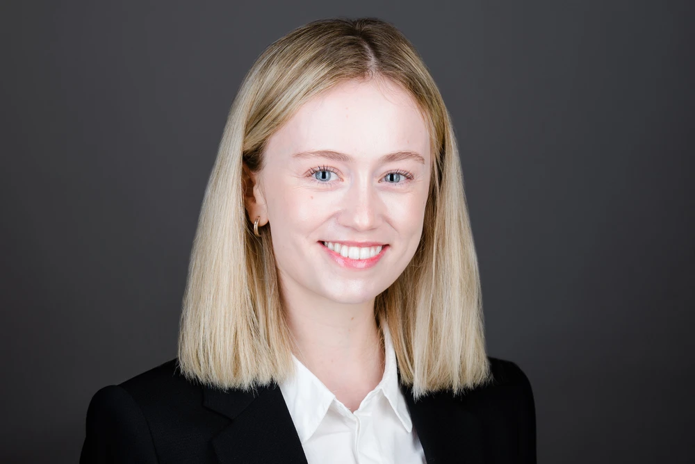 Smiling young woman with straight blonde hair, wearing a white shirt and black blazer, against a gray background.