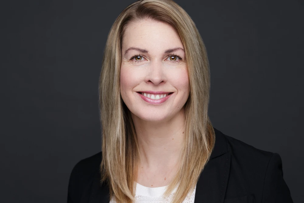 Smiling woman with straight blonde hair wearing a black blazer and white top against a dark gray background.