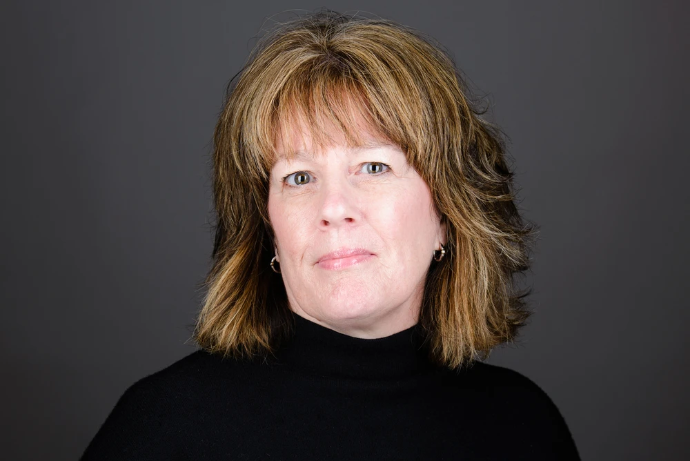Middle-aged woman with shoulder-length light brown hair wearing a black turtleneck against a dark gray background.